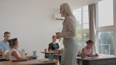 The-teacher-walks-around-the-classroom-of-the-school.-A-group-of-children-is-sitting-at-their-desks.-Children-write-down-the-task.-An-elementary-school-lesson-in-slow-motion.-Perform-control-work